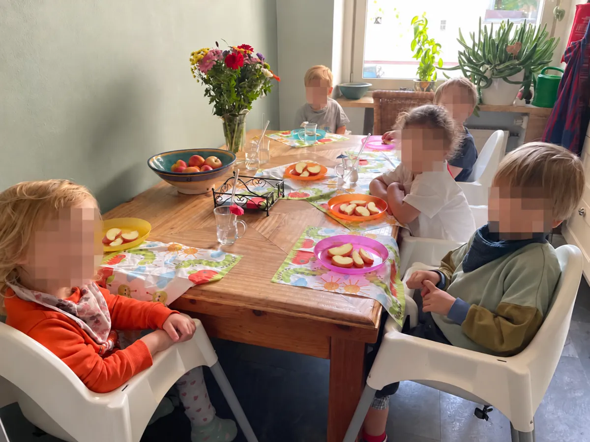 Fünf Kinder sitzen in Hochstühlen am Holztisch in Irenes Küche, vor jedem ein Teller mit Apfelstücken