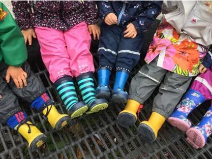 Kinder in Matschhosen und bunten Gummistiefeln sitzen nebeneinander draußen