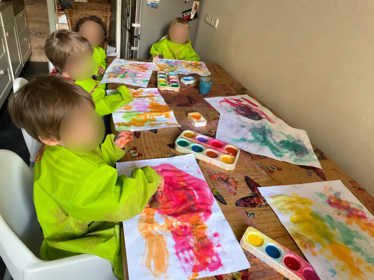 Kinder in grünen Malkitteln malen mit Wasserfarben am großen Küchentisch